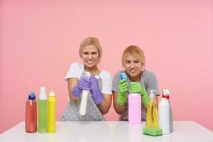 Indoor shot of young confused blonde cleaning women holding spray bottles with household chemicals frowning their faces while looking at camera, isolated over pink background photo