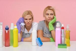 Indoor shot of tired young blonde cleaning ladies having hard working day and sitting at table with bottles of household chemicals, isolated over pink background photo