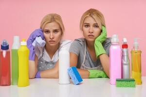 Portrait of young pretty white-headed cleaning ladies sitting over cleaning bottles and looking tiredly at camera, isolated over pink background in working clothes photo