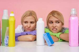 Aweary young brown-eyed blonde cleaning ladies holding their hands on countertop and looking tiredly at bottles with household chemicals, isolated over pink background photo