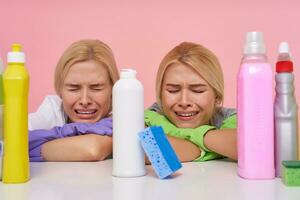 Unhappy young blonde cleaning ladies keeping their eyes closed while crying and holding head on folded hands, having too much work and being very tired, sitting over pink background photo