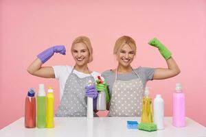 Indoor shot of young cheerful blonde cleaning women raising their hands while demonstrating power, being pleased with thier work, isolated over pink background photo