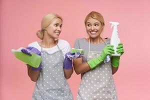 Studio photo of pretty young white-headed females keeping bottles with household chemicals and looking excitedly at camera. Housework and housekeeping concept