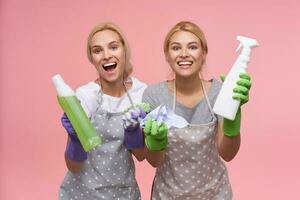 Cheerful young white-headed housewives in rubber gloves looking happily at camera with broad smiles and keeping cleaning stuff in raised hands, standing over pink background photo