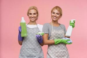 Happy young lovely blonde sisters with natural makeup holding bottles of household chemicals in raised hands and looking cheerfully at camera with broad smile, isolated over pink background photo