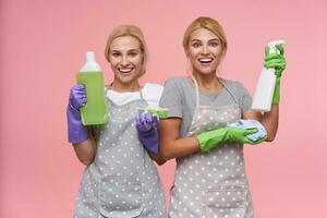 Joyful young white-headed cleaning ladies dressed in uniform keeping cleaning stuff in their hands while standing over pink background and smiling cheerfully photo