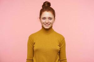 Portrait of attractive young redhead female with natural makeup looking positively at camera with charming smile, dressed in mustard sweater while posing over pink background photo