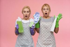 Astonished young blonde women in rubber gloves looking at camera with wide eyes and mouth opened while standing over pink background with detergents in raised hands photo