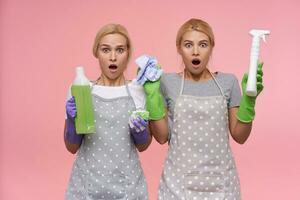 Dazed young pretty blonde cleaning ladies wearing casual t-shirts and polka dot apron looking amazedly at camera with opened mouths, isolated over pink background photo