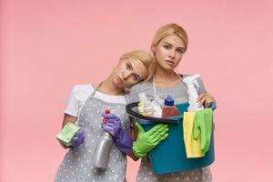 Indoor shot of young pretty blonde cleaning ladies holding detergents in raised hands and looking tiredly at camera, posing over pink background in working clothes photo