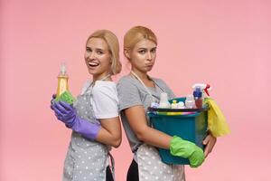 Studio photo of pretty young blonde cleaning ladies leaning back to back and expressing different emotions, keeping household chemicals in their hands while posing over pink background