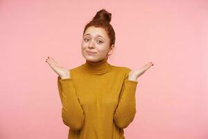 Young lady, pretty woman with ginger hair, freckles and bun. Wearing gold turtleneck sweater and shows with her posture misunderstanding. Watching at the camera isolated over pastel pink background photo