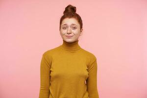 Adolescente chica, contento con su yo pelirrojo mujer con pecas y bollo. vistiendo oro suéter tipo con cuello de tortuga suéter, tener un linda sonrisa y acecho a el cámara aislado terminado pastel rosado antecedentes foto