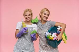 Happy young pretty blonde sisters with natural makeup rejoicing while finishing spring cleaning their house and posing over pink background with household chemicals photo