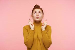 Desperate young lovely redhead female with bun hairstyle crossing her fingers while looking upwards with hope and pursing her lips, standing over pink background photo