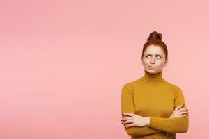 Teenage girl, wonder looking redhead woman with freckles and bun. Wearing gold turtleneck sweater and holds her arms crossed. Watching to the left at copy space over pastel pink background photo