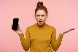 Studio photo of green-eyed young displeased redhead female raising perplexedly palm and frowning eyebrows while looking confusedly at camera, isolated over pink background