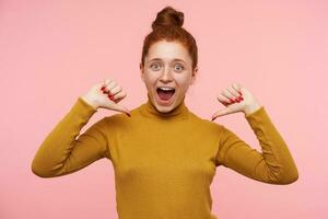 Teenage girl, happy looking redhead woman with freckles and bun. Wearing gold turtleneck sweater and pointing at her self with thumbs. Watching at the camera isolated over pastel pink background photo