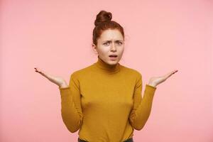 Puzzled young green-eyed redhead lady with natural makeup frowning her eyebrows and raising confusedly palms while looking at camera, standing over pink background photo