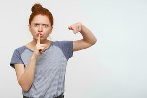 Serious young pretty redhead female with bun hairstyle pointing at camera with raised forefinger and raising hand with hush gesture, posing over pink background photo