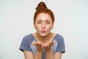 Positive young redhead female with natural makeup folding her lips and blowing air kiss at camera, keeping palms raised while posing over pink background photo