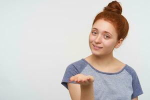 Positive young pretty redhead lady with bun hairstyle keeping her palm raised and looking at camera with gentle smile, standing over pink background in casual clothes photo