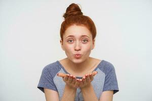 Studio shot of young lovely positive redhead lady keeping her palms raised and blowing air kiss at camera, wearing casual clothes while posing over pink background photo
