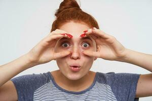 Close-up of excited young redhead female with red manicure looking at camera with wide eyes opened and fooling while standing over pink background in casual wear photo