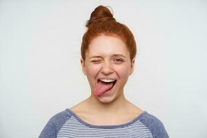 Joyful young loely redhead woman with natural makeup winking at camera and sticking happily out her tongue, having fun while posing over pink background photo