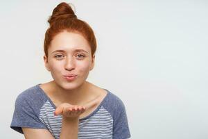 Portrait of young pretty redhead lady with bun hairstyle folding her lips in air kiss and raising hand while looking positively at camera, isolated over pink background photo