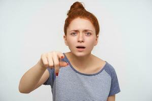 Puzzled young attractive redhead lady frowning her eyebrows while looking at camera and pointing ahead with raised forefinger, isolated over pink background photo