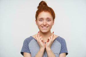 Hoping young positive redhead woman with natural makeup biting underlip and crossing her fingers with red manicure while making wish, standing over pink background photo