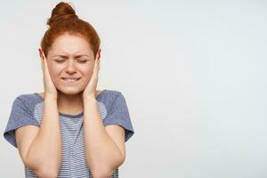 Studio photo of young displeased redhead female keeping her eyes closed and covering ears with raised palms while avoiding loud sounds, isolated over pink background