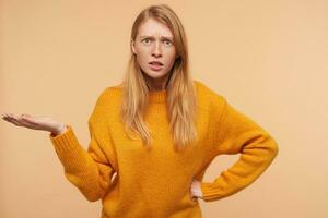 Confused young pretty redhead female frowning her eyebrows and raising perplexedly palm while standing over beige background in knitted mustard pullover photo