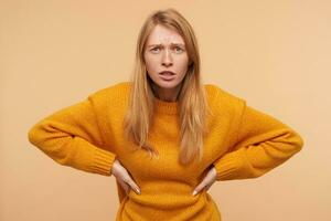 Bewildered young pretty long haired redhead lady keeping hands on her waist while looking confusedly at camera and squinting her green eyes, isolated over beige background photo
