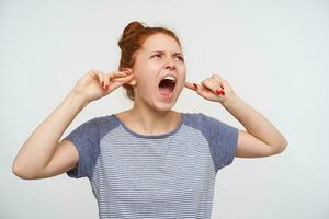 Stressed young angry redhead lady frowning her face while screaming crossly and inserting forefingers into ears because of annoying sounds, standing over pink background photo