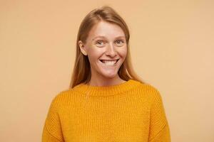 Portrait of young attractive glad lady with loose foxy hair looking at camera with charming smile and biting her underlip, standing against beige background photo