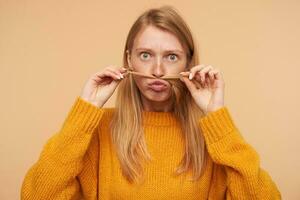 Funny shot of young green-eyed redhead woman imitating mustache with lock of hair and looking excitedly at camera, isolated over beige background in mustard sweater photo