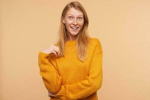 Surprised young attractive redhead woman with casual hairstyle touching her hair and looking emotionally at camera, posing over beige background in mustard sweater photo
