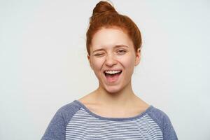 Joyous young lovely redhead female with bun hairstyle winking happily at camera and smiling widely, wearing striped blue t-shirt while posing over pink background photo