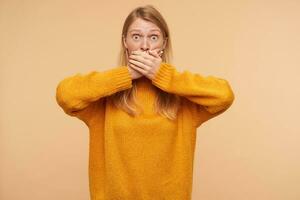 Amazed young pretty redhead woman with casual hairstyle covering her mouth with raised hands while looking dazedly at camera, standing over beige background photo