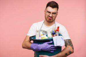 Disgruntled young short haired brunette male keeping busket with detergents and frowning his face while looking at camera, having spring cleaning on weekend and beeing upset about it photo