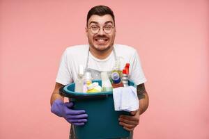 Positive young handsome dark haired male in rubber glove looking excitedly at camera and smiling cheerfully, standing over pink background in working clothes photo