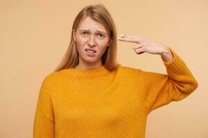 Displeased young pretty redhead female with casual hairstyle looking at camera with pout and folding gun with raised hand, standing over beige background photo