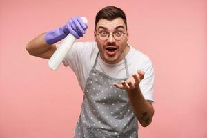 Excited young pretty dark haired guy in eyeglasses raising hand with white spray bottle and looking at camera with wide eyes and mouth opened, isolated over pink background photo