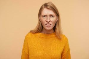 Dissatisfied young attractive long haired redhead female frowning discontentedly her face while looking at camera, dressed in casual clothes while posing over beige background photo