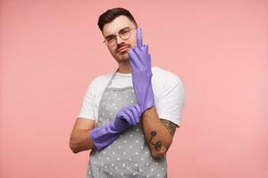 Cocky young short haired brunette male in eyeglasses putting on rubber gloves and showing raised middle finger, wearing basic white t-shirt and apron while posing over pink background photo