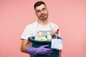 Displeased young brunette male with short haircut grimacing his face while keeping busket with bottles of household chemicals, standing over pink background in uniform photo