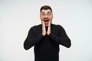 Bemused young pretty dark haired bearded guy holding palms on his face while looking amazedly at camera with wide eyes and mouth opened, isolated over white background photo