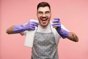 Agitated young pretty brunette male with short haircut looking excitedly at camera while holding spray bottle in raised hand, posing over pink background photo